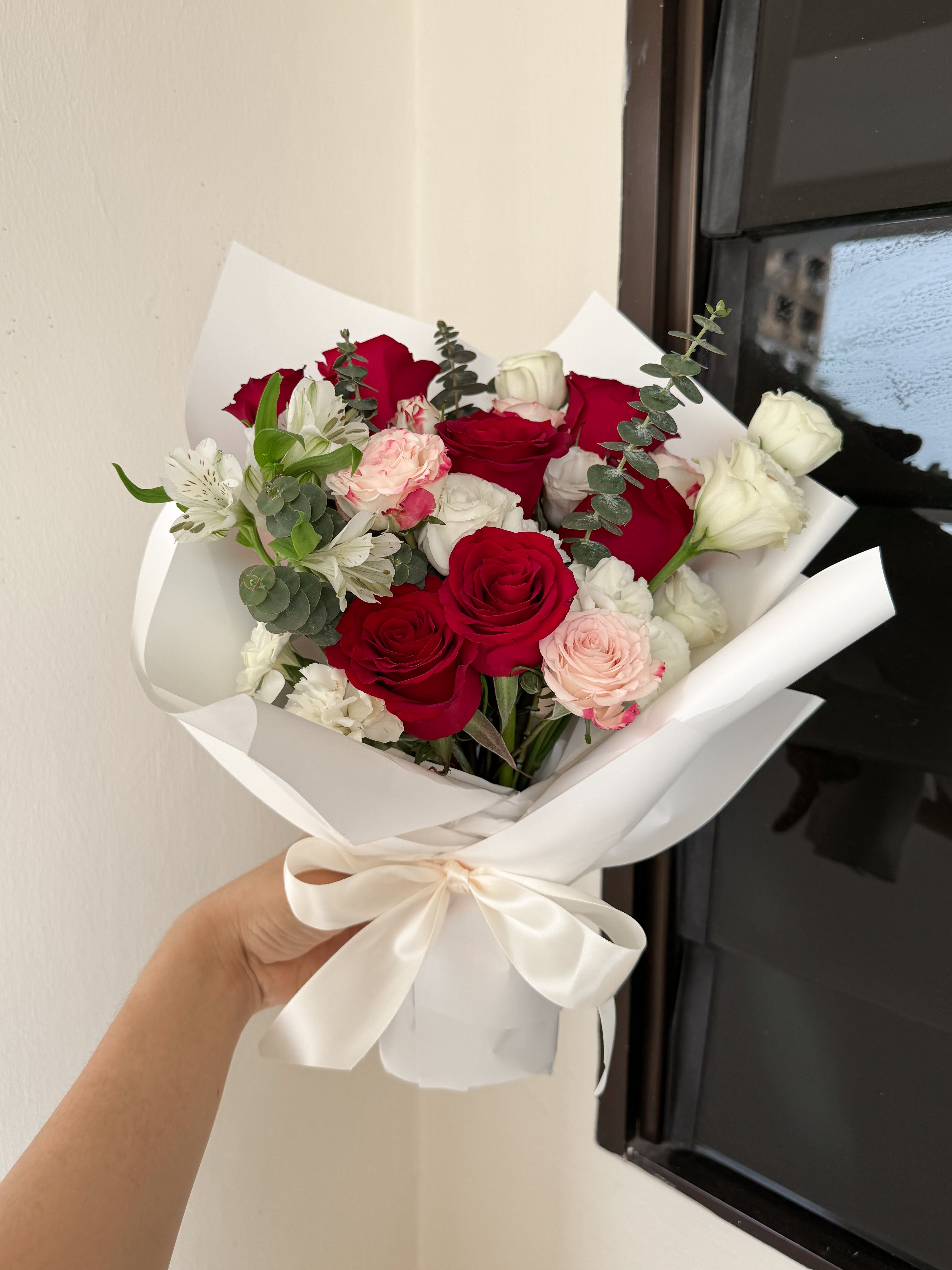 Bouquet of red, white, and pink roses wrapped in white paper with a bow, held against a neutral background.